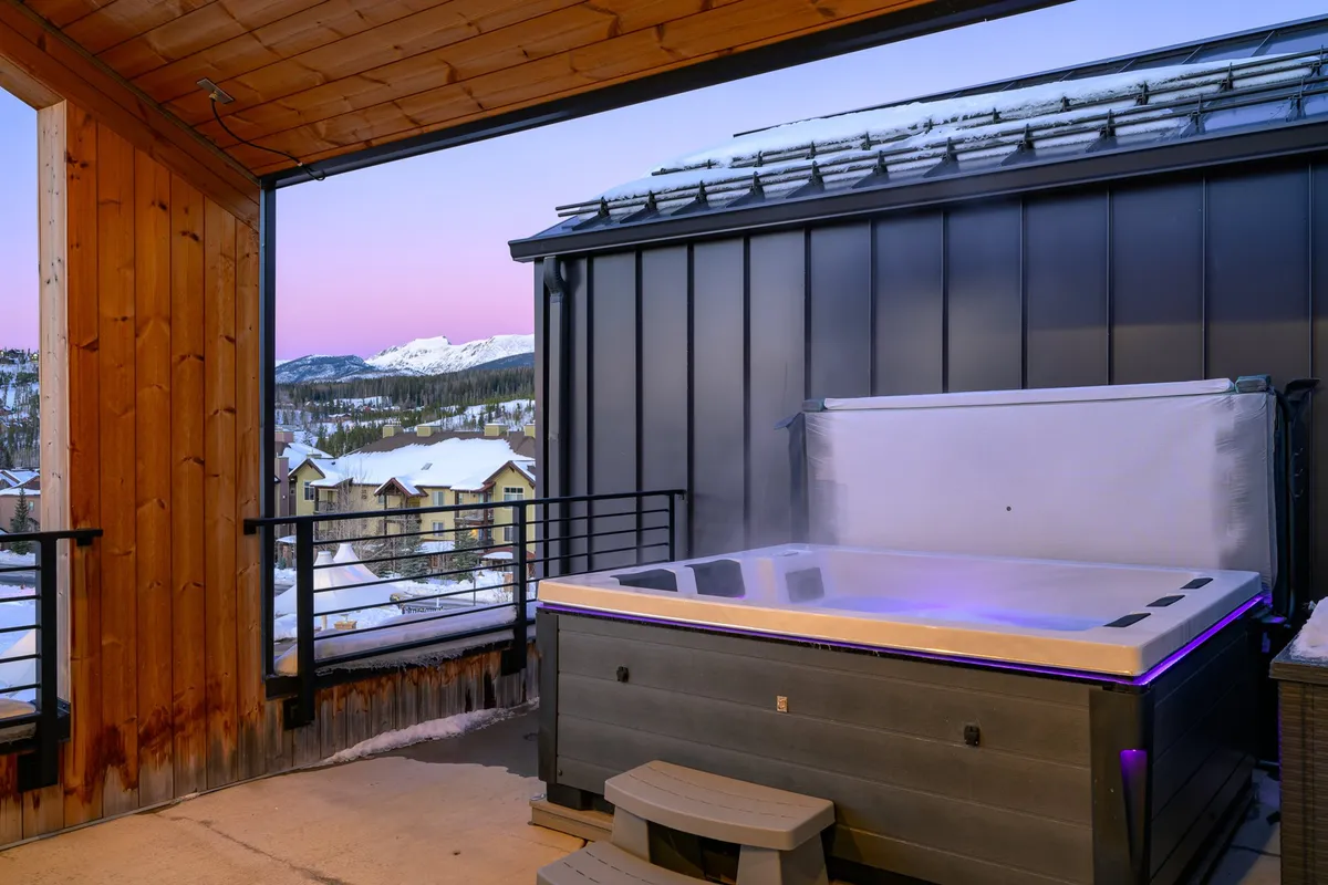 A tranquil exterior rooftop scene featuring a bubbling hot tub with sweeping views of the mountains, framed by wooden beams and railings. The warm water of the hot tub contrasts with the cool mountain air, and the sky glows with the colors of sunset, enhancing the relaxing ambiance. Lounge seating is positioned nearby, inviting guests to unwind and enjoy the vista.