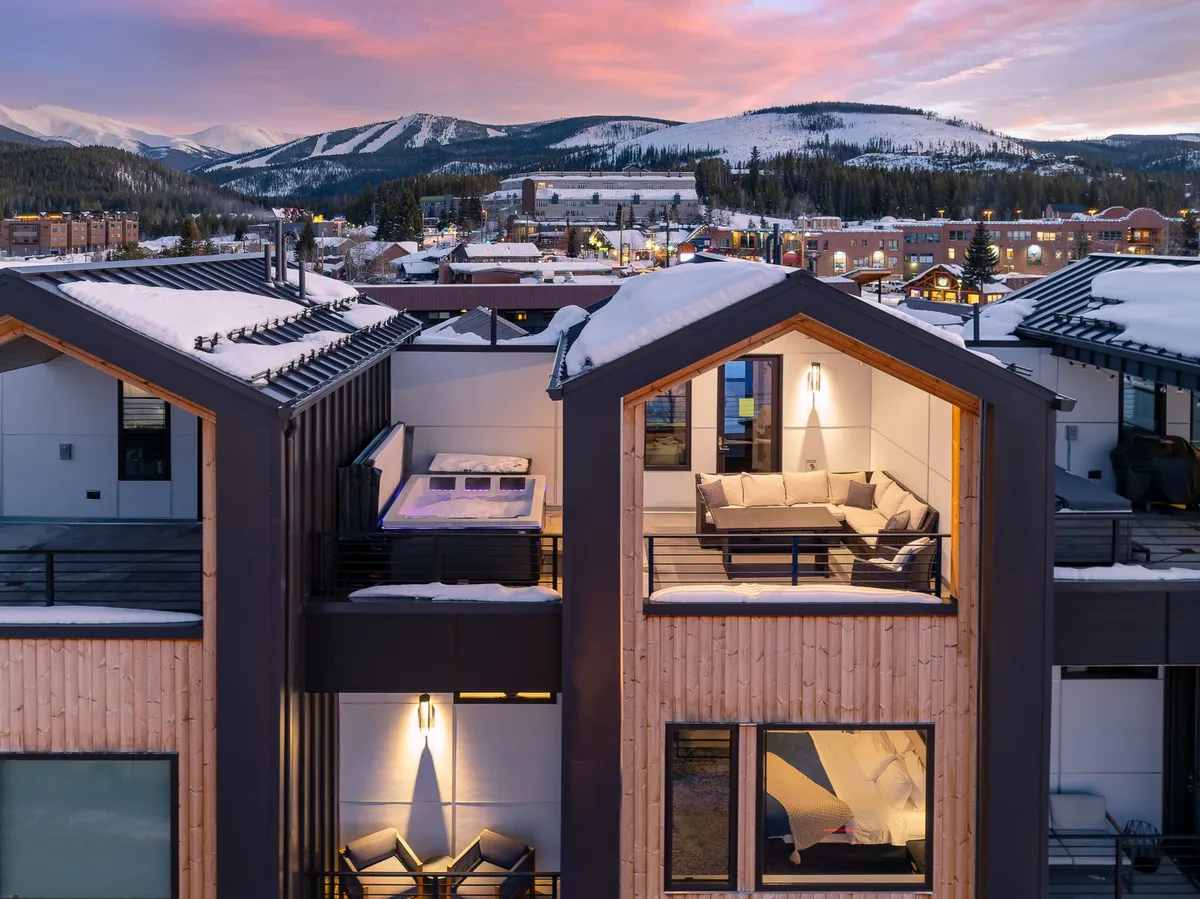 A festive rooftop patio scene with modern architecture, snow-covered roofs, a bubbling hot tub, and a spacious outdoor seating area. The setting is lively but tranquil, inviting guests to immerse themselves in the hot tub while snow gently falls, or gather around the fire pit for conversation and warmth. The space offers direct sightlines to the vibrant life of Winter Park below.