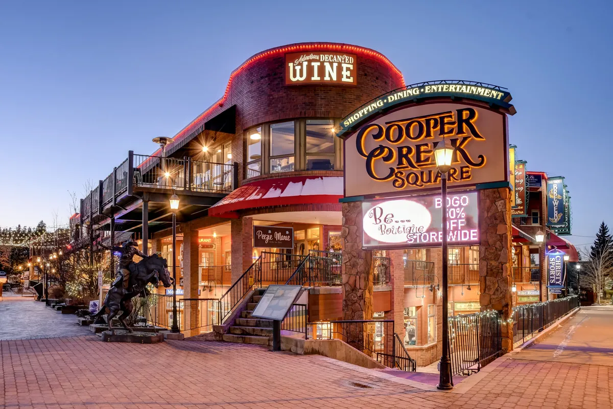 A festive evening scene in Cooper Creek Square, featuring the illuminated sign, glowing storefronts, decorative lighting, and a statue out front. Shoppers and diners add energy, highlighting the area’s vibrant culture and entertainment offerings.
