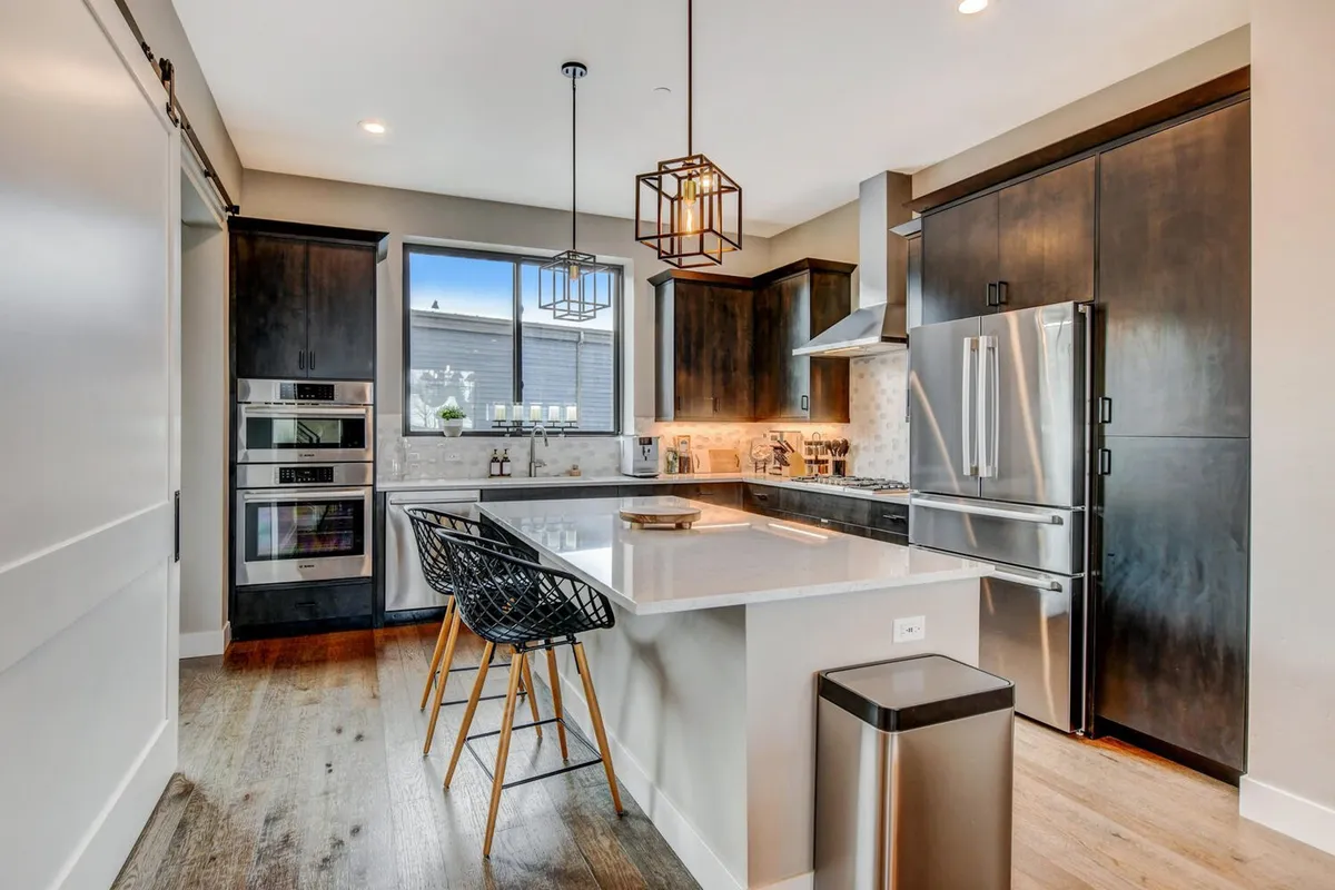 A contemporary kitchen with a sleek island at its center, surrounded by stainless steel appliances, elegant pendant lighting, and streaming natural light. The setup is ideal for cooking, hosting, and casual conversations, reflecting a high-end, functional kitchen space ready for any culinary endeavor.