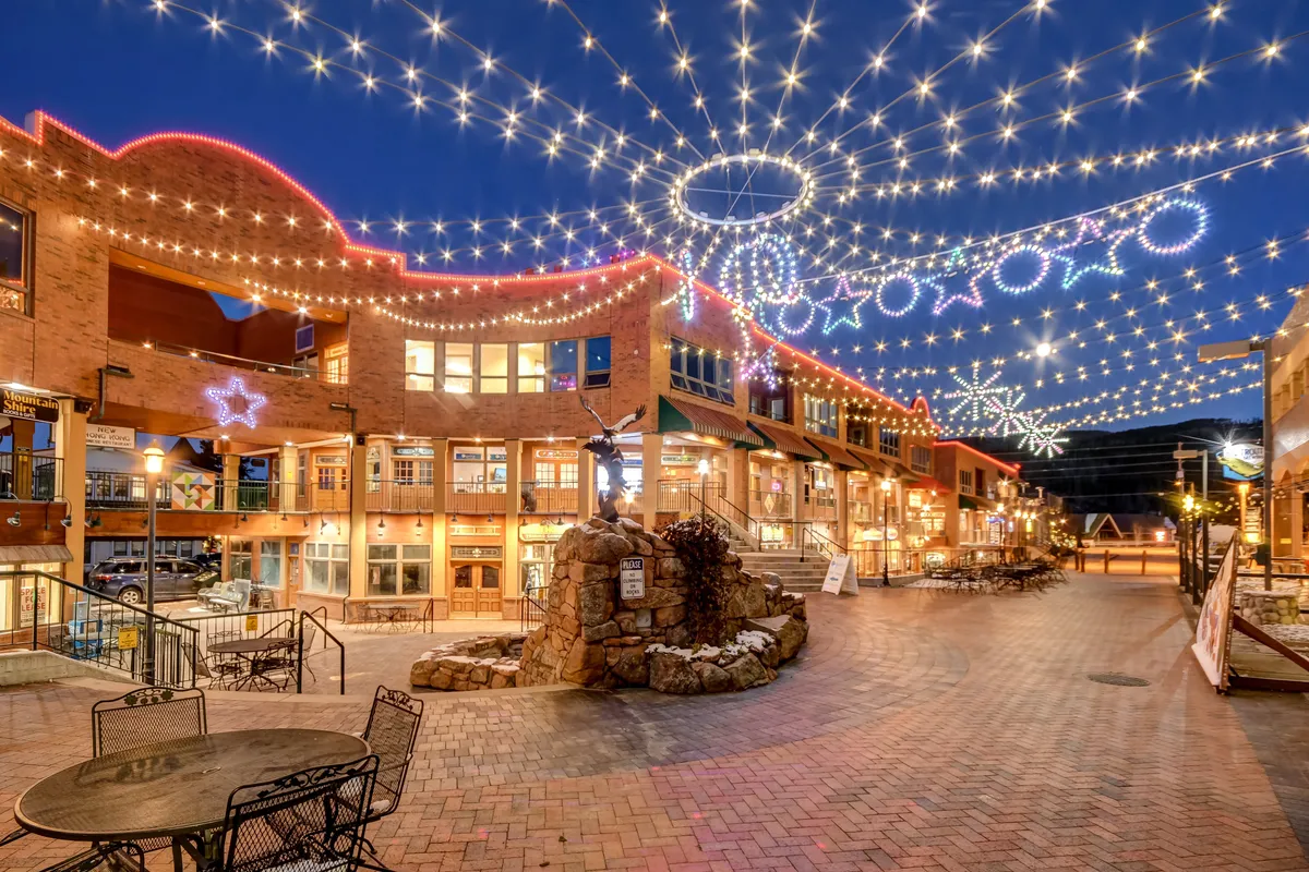 A vibrant streetscape with string lights, decorative stars, a brick pathway, storefronts, and a fountain, capturing the festive, social atmosphere of the local neighborhood. The scene suggests walkable access to dining, shopping, and community activities.