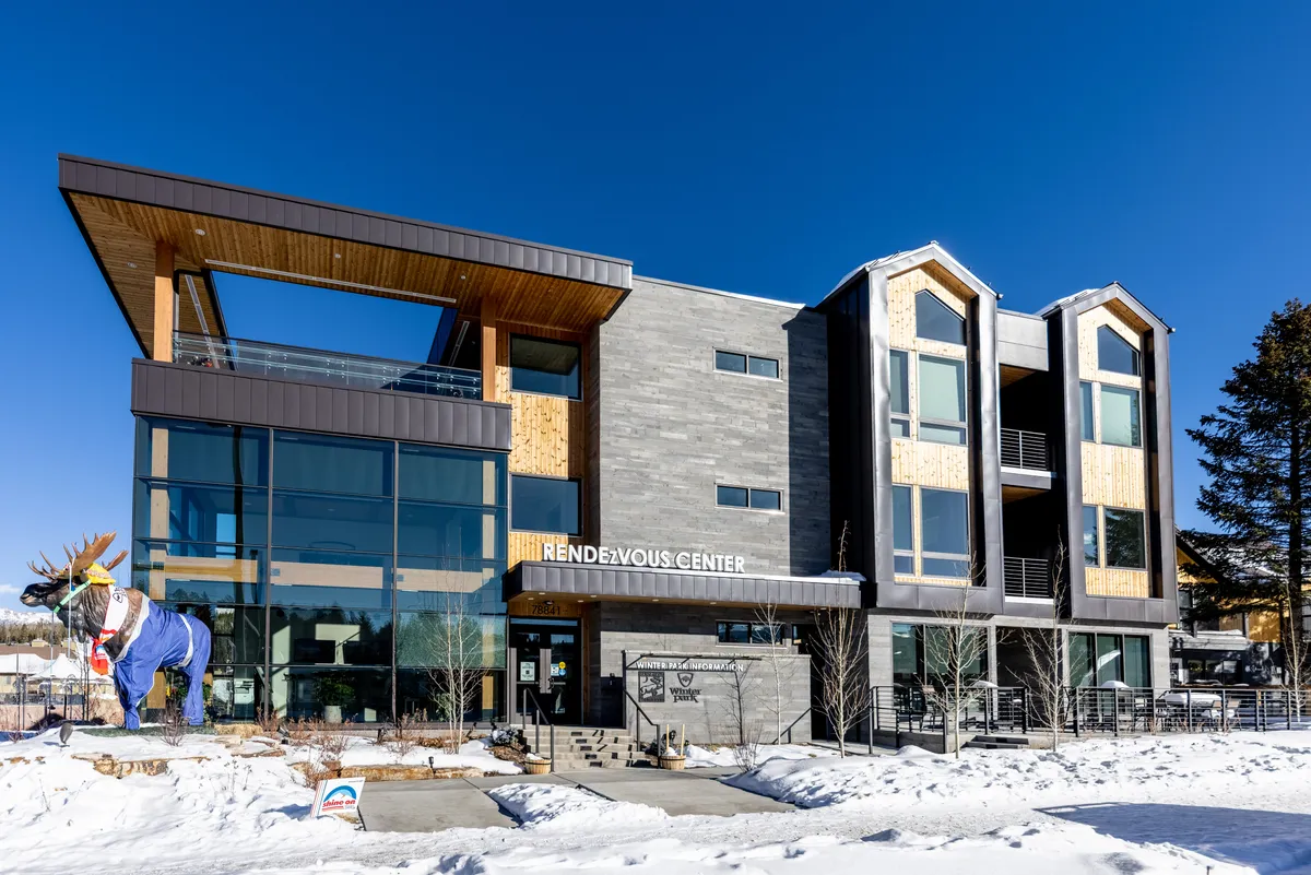 A striking exterior view of the building showcasing modern architecture, expansive windows, and a snow-dusted setting. The scene includes a prominent statue of a moose and hints at communal gathering spaces outdoors, all set against a wintery backdrop.