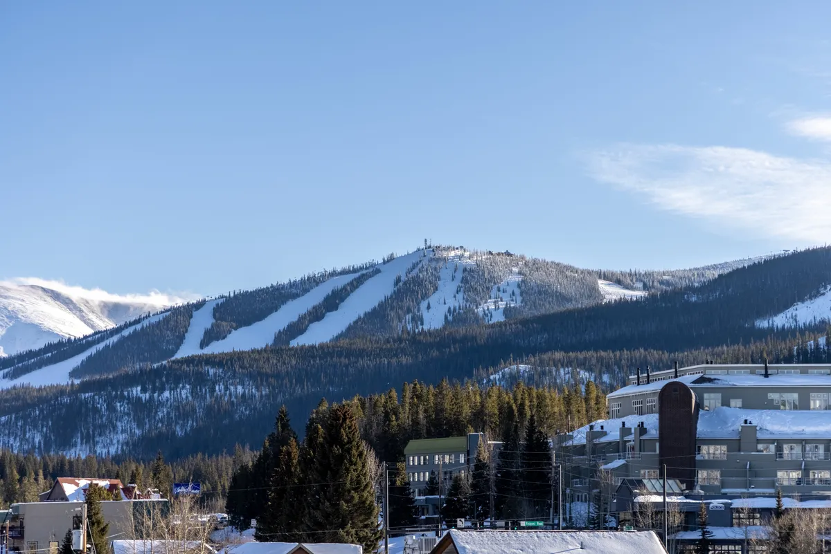 A breathtaking view of snow-covered mountains, ski slopes, and dense pine forests with nearby buildings in the foreground. The scene evokes adventure and the beauty of the natural surroundings, capturing the atmosphere of the mountain setting.