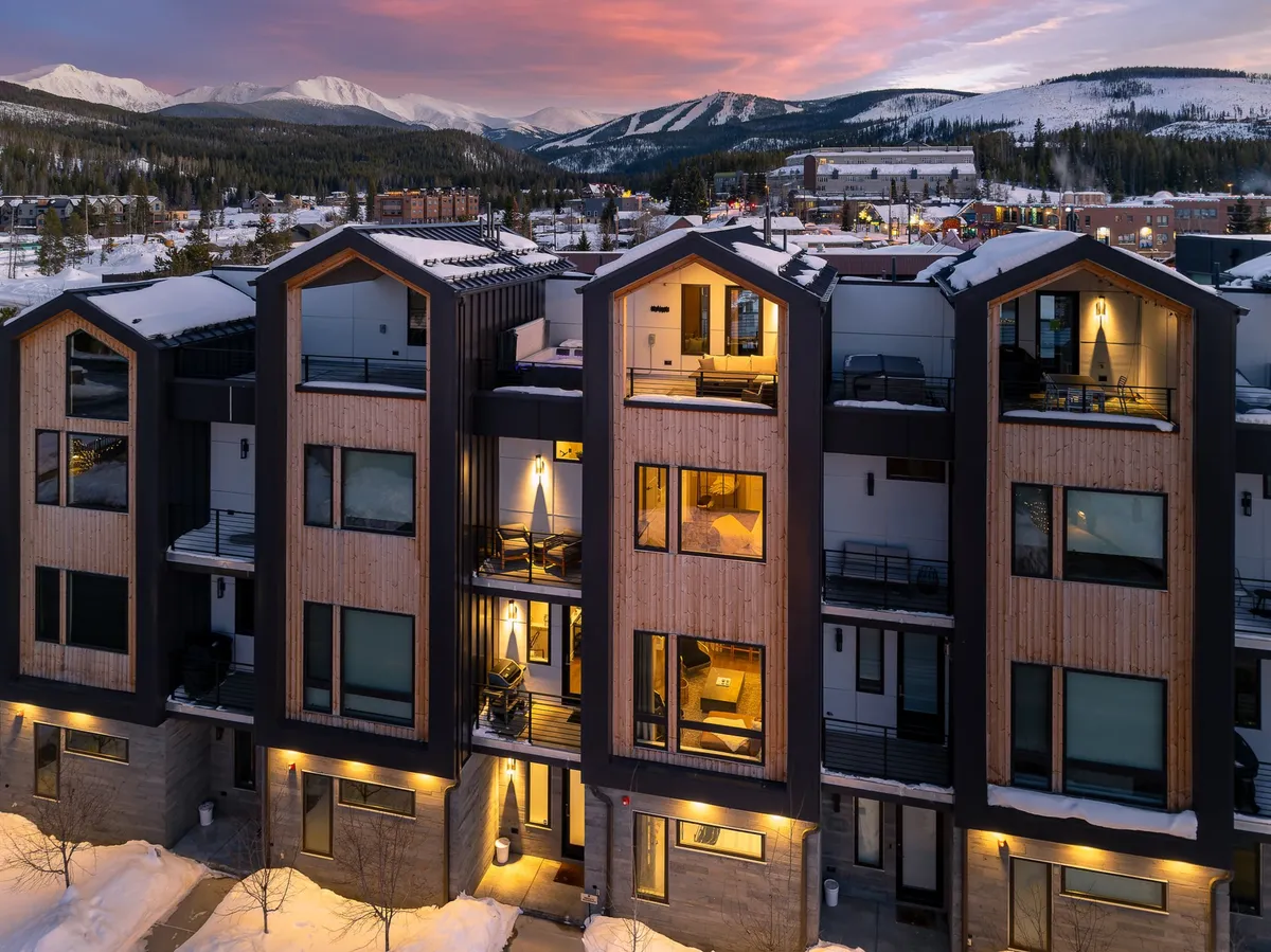 The exterior of a modern mountain home with sleek architectural lines, large windows, and a snow-covered landscape framed by towering peaks. The property exudes a sense of luxury, adventure, and refined comfort, while the surrounding natural beauty hints at both resort proximity and serene getaways.