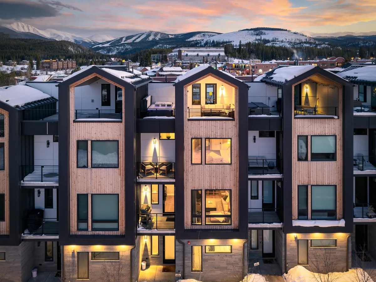 The image features a striking, modern single-family home set against a dramatic mountain backdrop at dusk. Warm golden light spills from the windows, highlighting inviting interiors after a day of adventure. The property’s balconies and outdoor spaces are perfect for socializing, all framed by breathtaking alpine views.