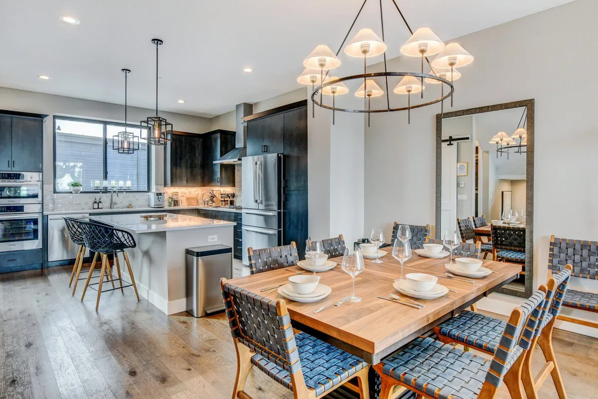 An inviting open-concept dining and kitchen area, with a large dining table set for a group, a kitchen island with bar seating, modern lighting fixtures, and ample natural light pouring through spacious windows. The space is designed for both festive feasts and casual breakfasts, seamlessly connecting cooking and dining zones.