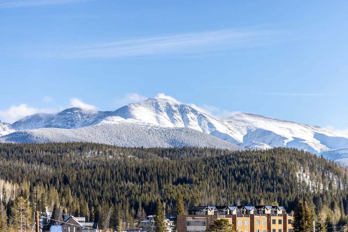 A stunning view of snow-capped Rocky Mountains rising above a dense evergreen forest, with a modern building in the foreground. The scene is tranquil yet inspiring, hinting at the proximity of top attractions and natural beauty available to explore right outside the property.