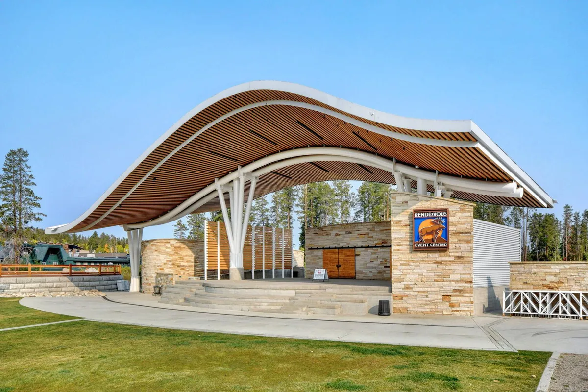 A modern outdoor stage with a curved roof and stone facade, surrounded by wooden beams and an inviting plaza space. The setting is designed for gatherings, concerts, and lively events under the open sky.
