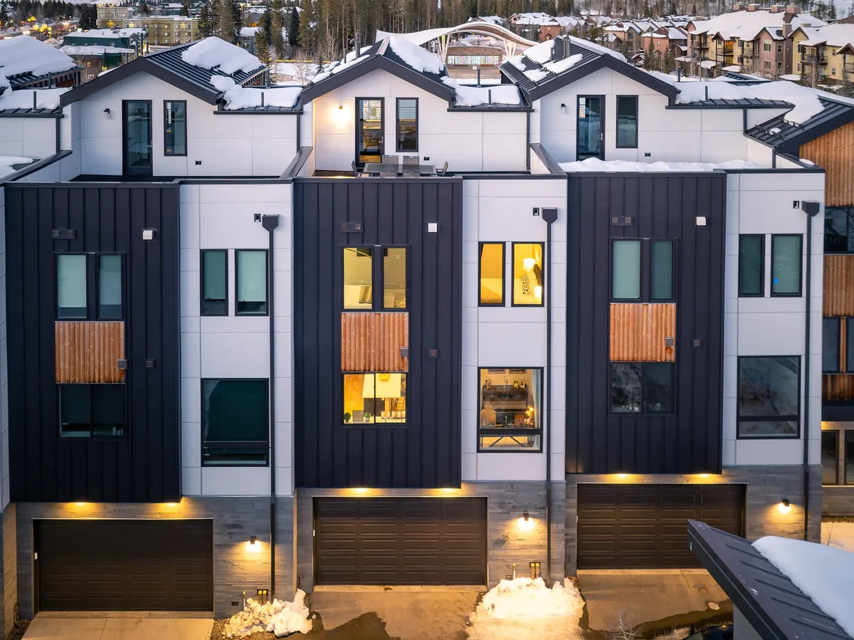 A contemporary exterior view of the home with modern architecture, symmetrical windows, wooden accents, and snow-covered roofs, evoking a welcoming and cozy atmosphere. The setting highlights the home's capacity for group stays and its inviting presence in a serene neighborhood.