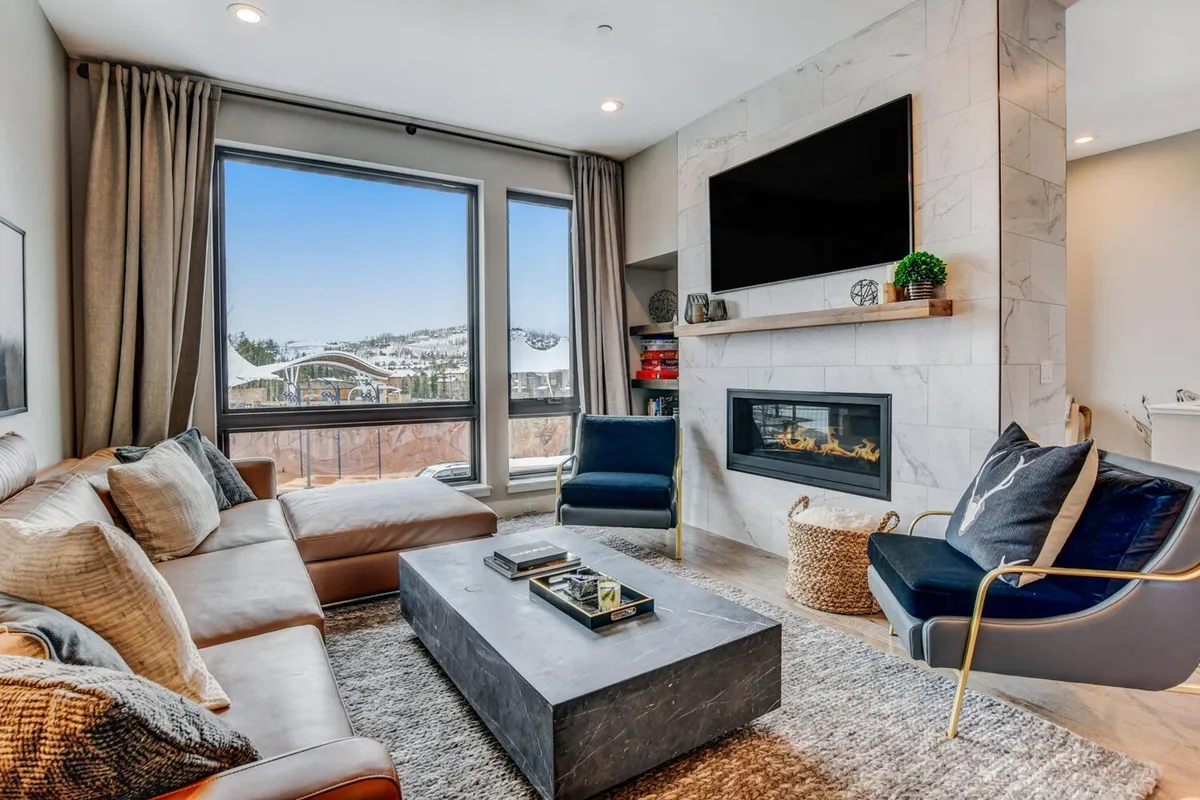 A spacious, sun-filled great room featuring expansive windows with mountain views, a modern fireplace, a plush sectional sofa, stylish accent chairs, and a decorative coffee table. The arrangement invites group relaxation, with direct sight lines to the outdoors and a cozy ambiance perfect for gatherings or movie nights.