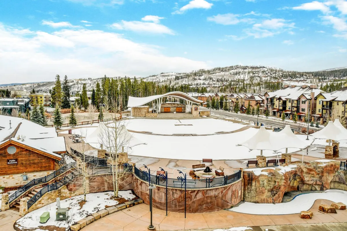 A vibrant winter scene at the amphitheater stage in Hideaway Park, with snow covering the ground, colorful buildings lining the background, and trees framing the area. The amphitheater is clearly central, suggesting its use for outdoor concerts and community events.