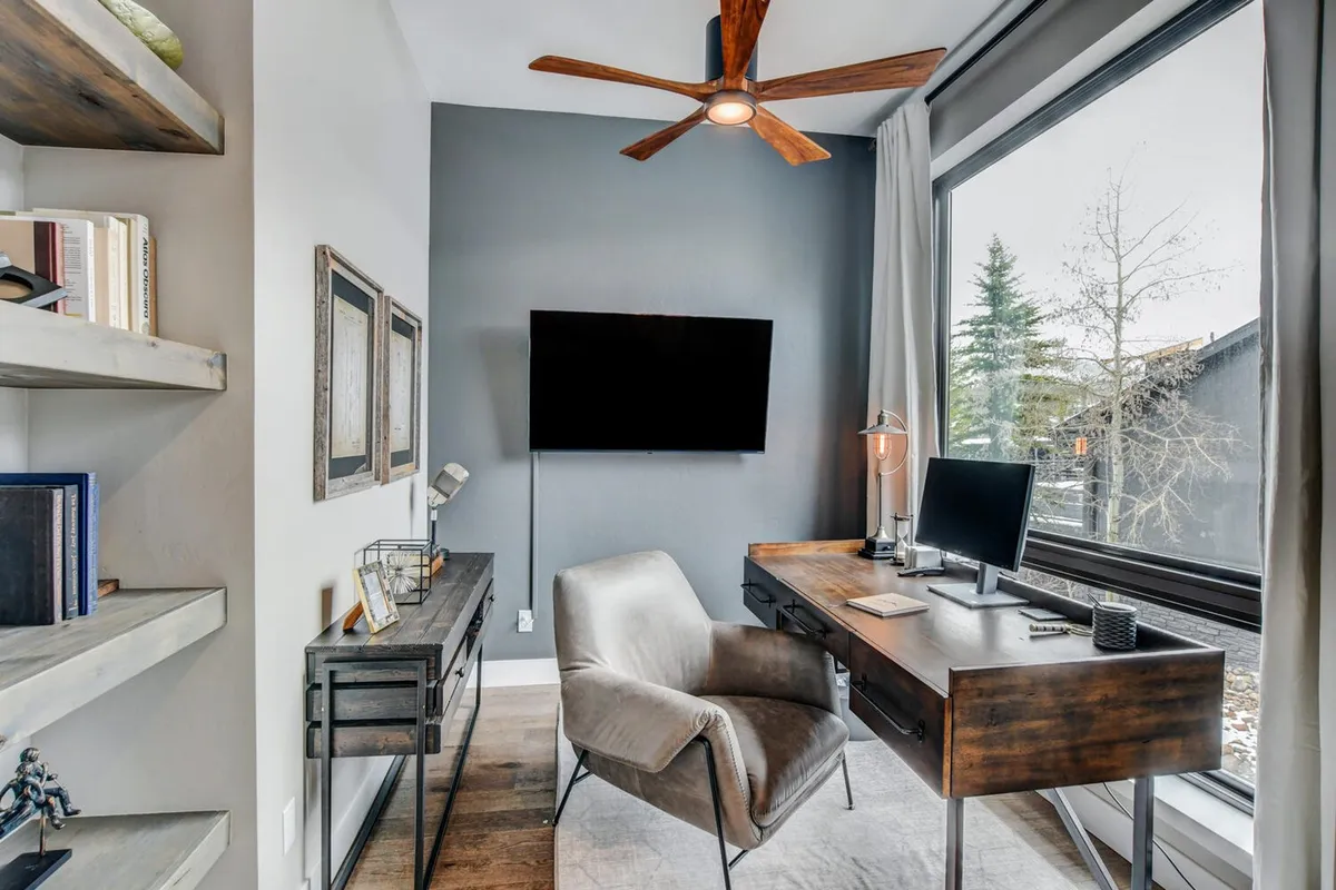 A private office suite with a wooden desk, comfortable chair, and a large window that lets in plenty of natural light. Shelves decorated with art and plants create a productive yet calming work environment, with views out onto the landscape.