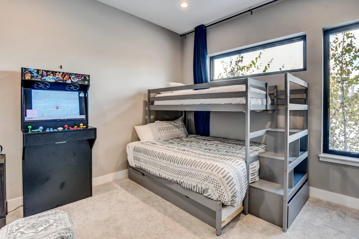 A fun and inviting bunkroom featuring a bunk bed, bright windows, and a large arcade game machine. The room is designed for both sleep and entertainment, with ample light and space for group activities.